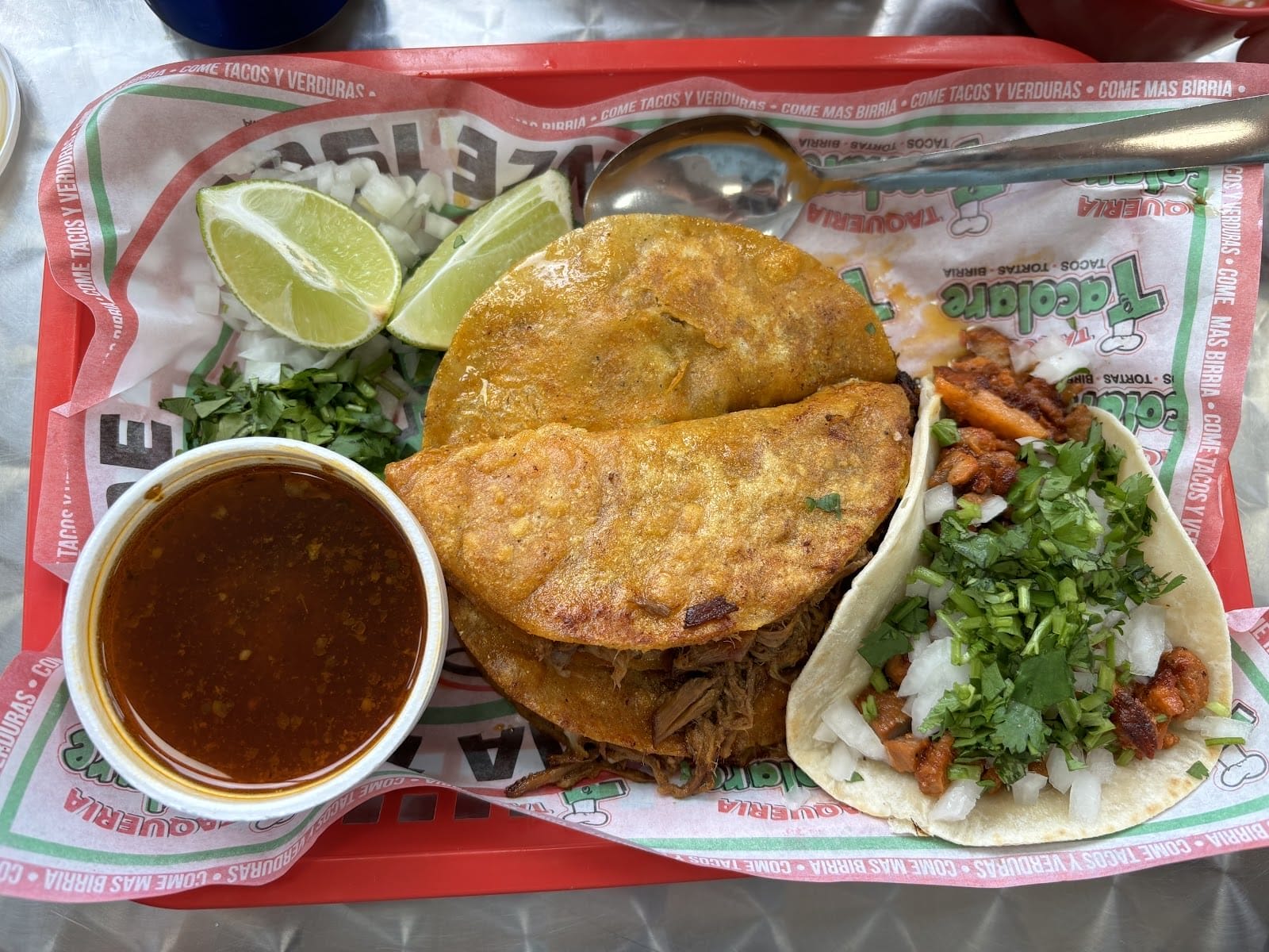 Ein Tablett mit mexikanischen Speisen, darunter zwei Birria-Tacos mit Brühe, gehackten Zwiebeln, Koriander, Limettenspalten und einem kleinen Taco. Das Gericht ist mit frischen Kräutern garniert, und ein Löffel wird neben das Essen gelegt.