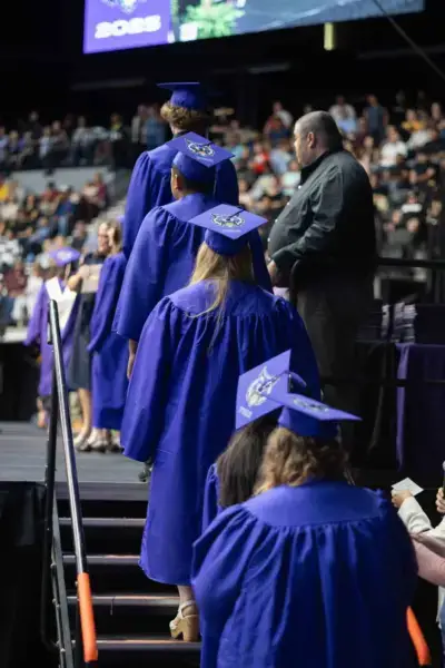 Absolventen in blauen Roben und mit geschmückten Mützen betreten eine Bühne bei einer Zeremonie. Im Hintergrund sieht eine Menschenmenge zu, die eine feierliche Atmosphäre schafft.