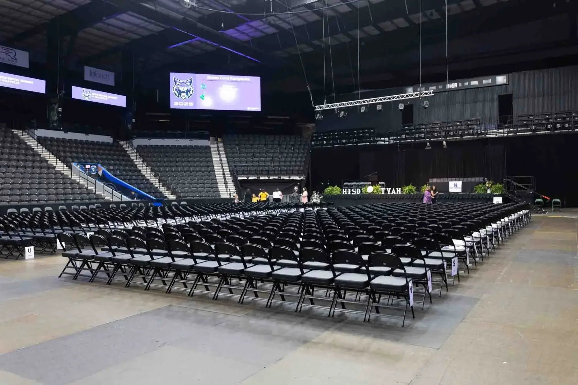 Eine leere Halle mit Reihen von schwarzen Stühlen vor einer Bühne. Auf großen Bildschirmen sind Logos zu sehen, und grüne Pflanzen schmücken die Bühne und schaffen eine erwartungsvolle Atmosphäre.